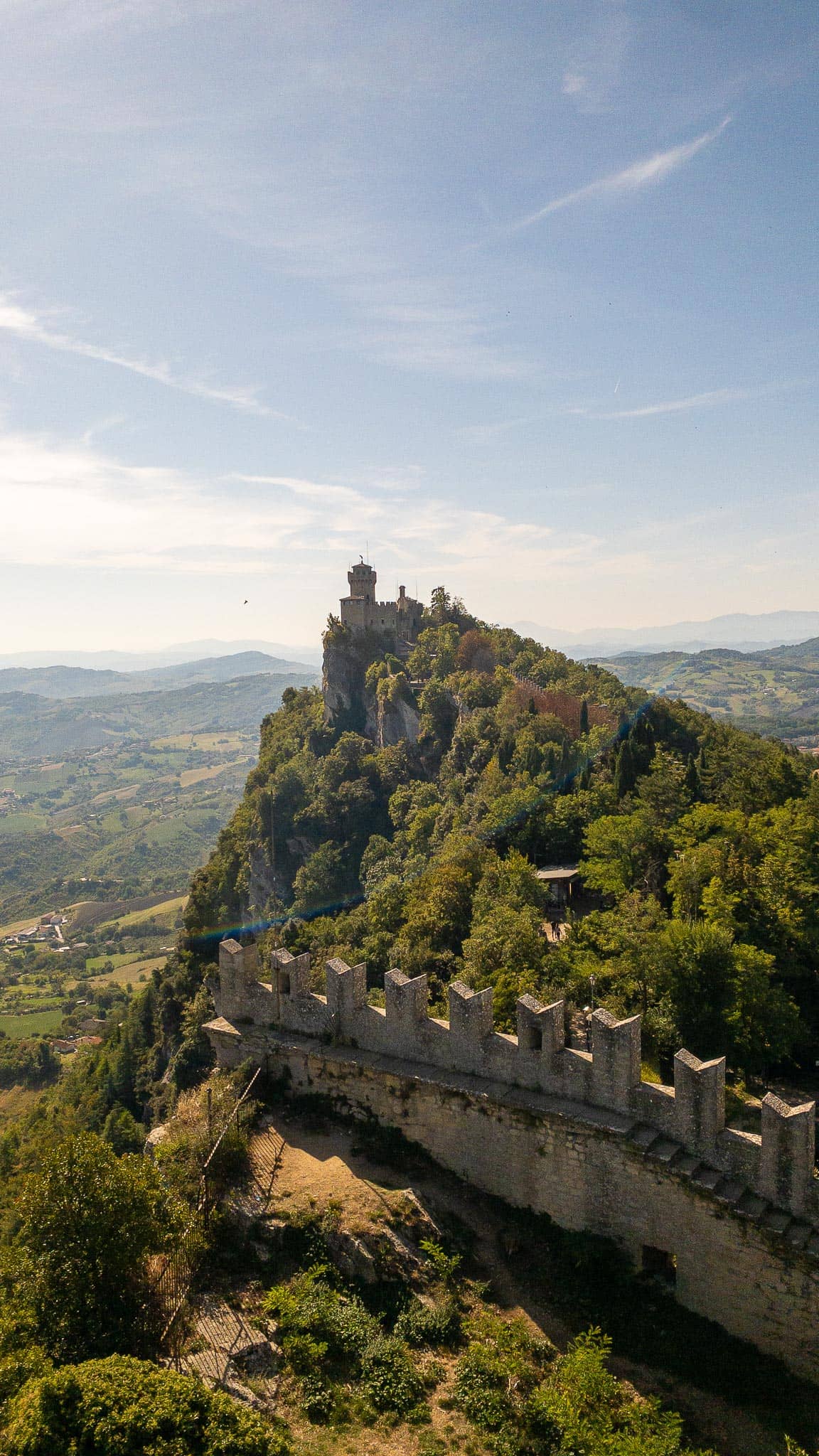 A crenellated rampart wall along the wooded ridge of Monte Titano in San Marino, with a stone tower ahead and the plain visible far below on the right.
