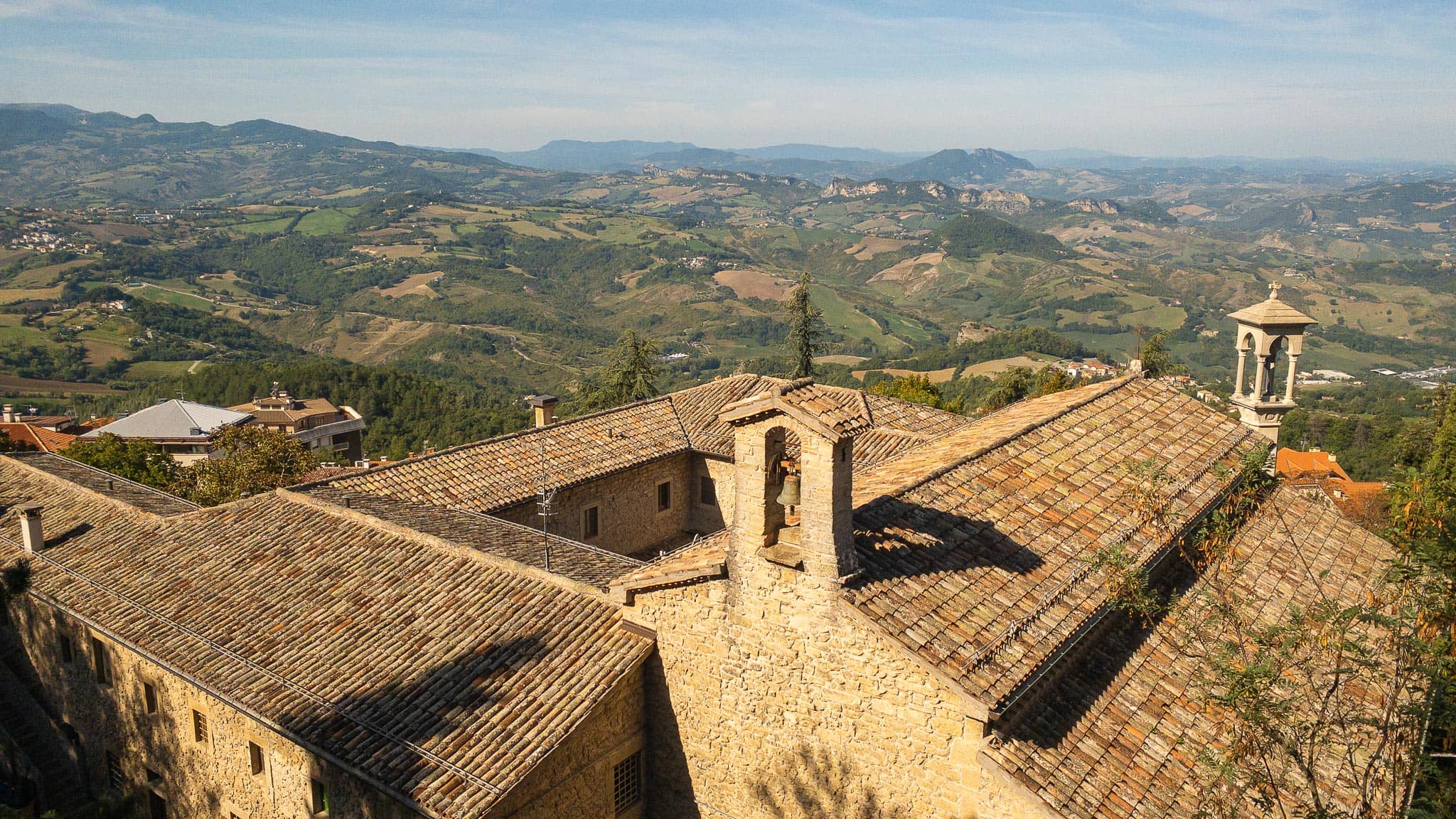 Stone buildings with pale tiled roofs and a small bell turret in San Marino, looking out over green farmland and low hills fading into haze.