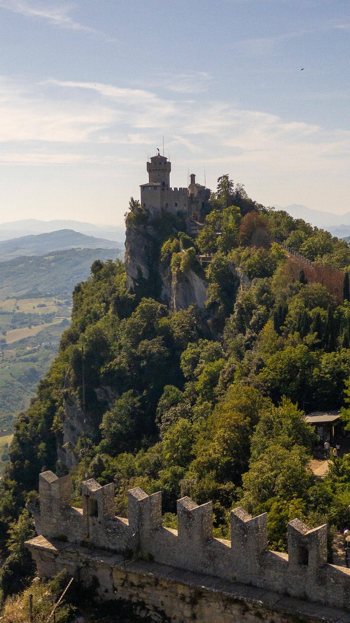 One of San Marino's stone towers standing on a wooded limestone crag, seen along a crenellated rampart wall running out towards it.