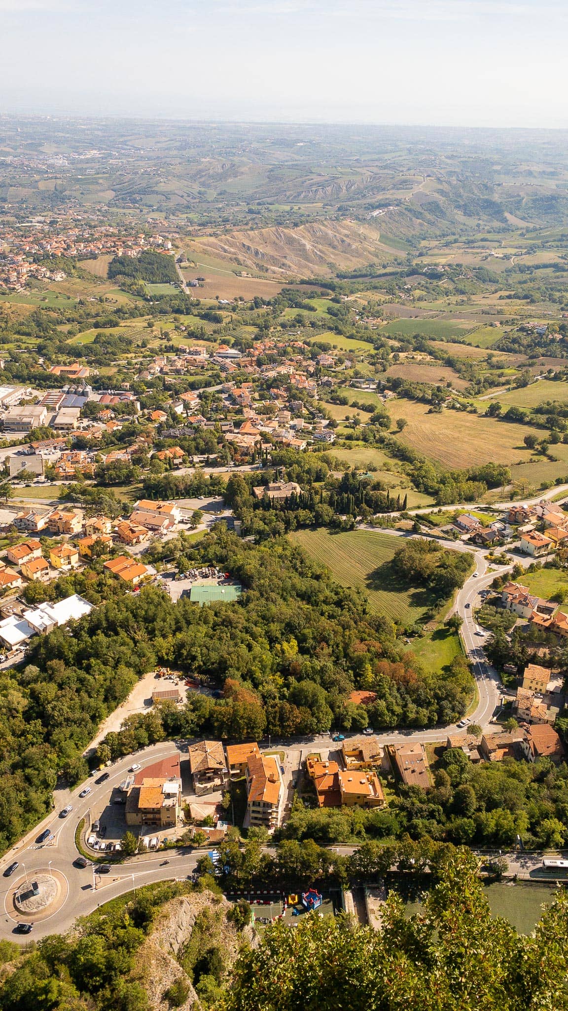 Looking down from Monte Titano over red-roofed houses, wooded slopes and a patchwork of fields and roads reaching to the horizon.