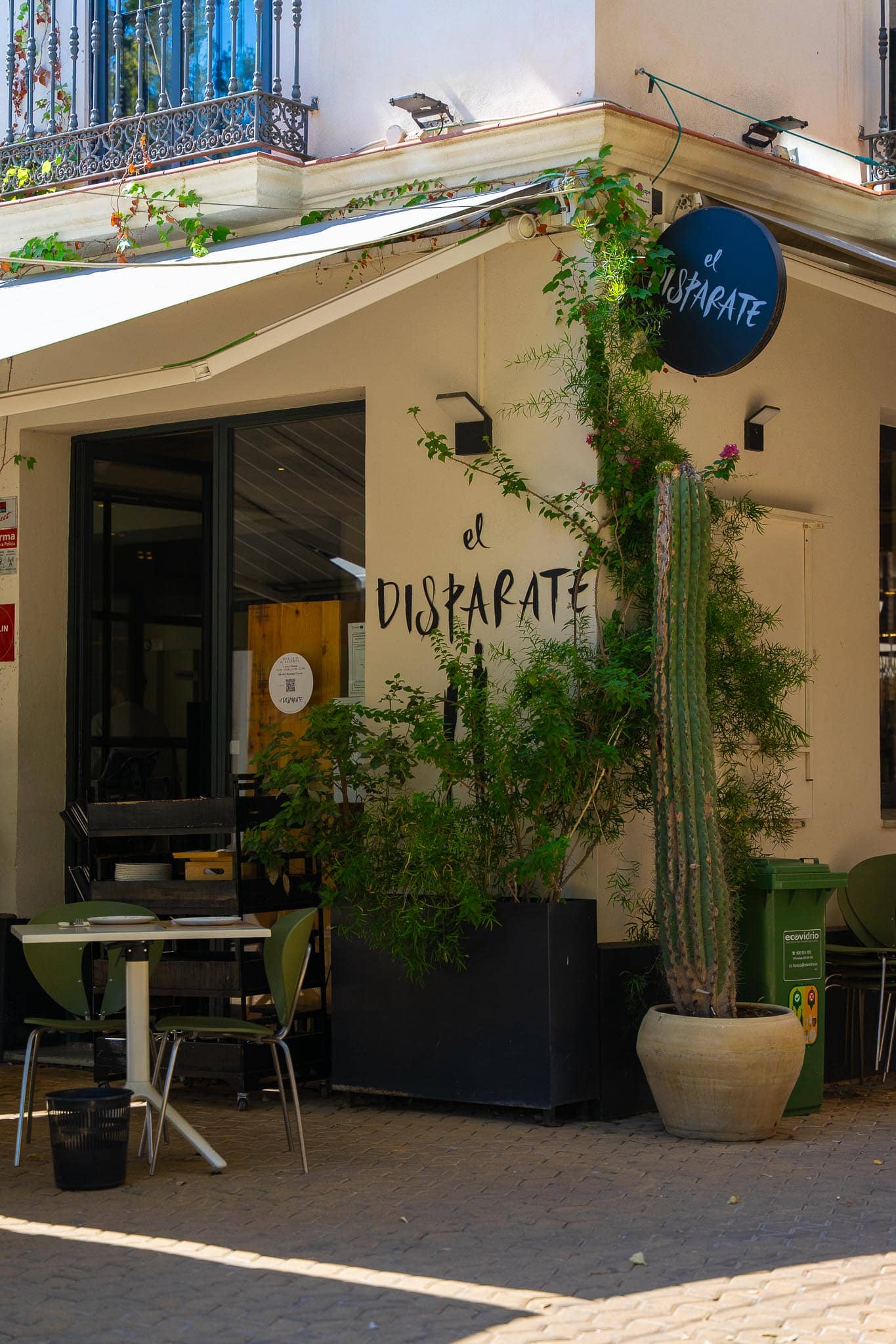 The corner exterior of a restaurant called el Disparate on the Alameda de Hércules in Seville: cream walls with the name hand-painted in large black brush lettering, a round navy sign above, a tall cactus in a terracotta pot, bougainvillea trained up the wall, and one laid pavement table with green chairs.