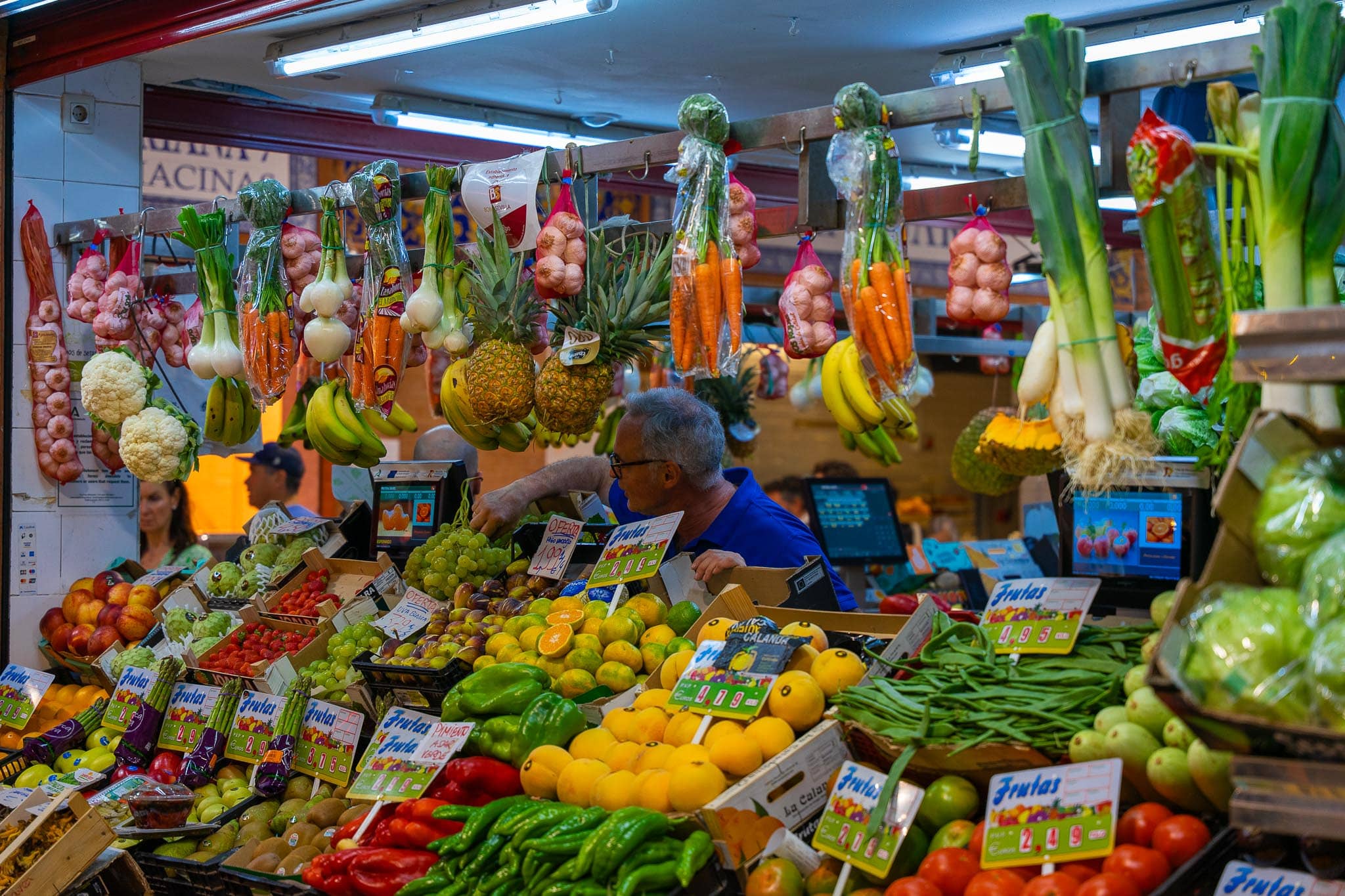 A greengrocer's stall in the Mercado de Triana: bunches of garlic, spring onions, carrots, leeks, pineapples, bananas and cauliflowers hanging from a rail above crates of lemons, oranges, peppers, green beans and strawberries, with a stallholder in a blue polo shirt reaching across and handwritten Frutas price cards throughout.