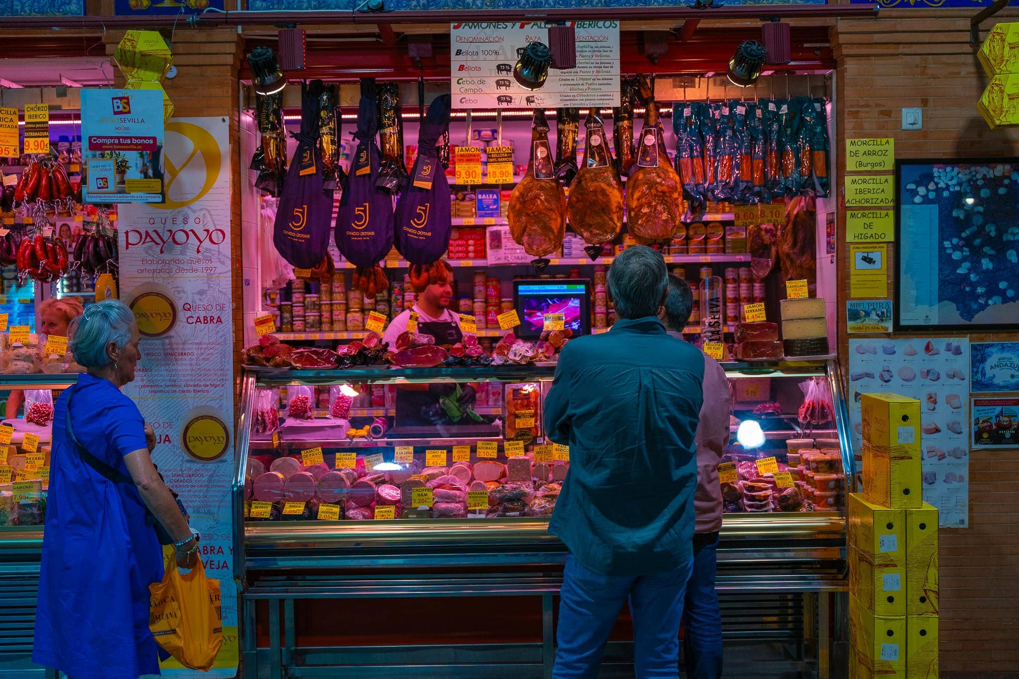 A charcuterie counter in the Mercado de Triana under magenta lighting: whole Ibérico hams hanging in dark netted sleeves and unwrapped, rings of chorizo and morcilla, a Queso Payoyo poster, a printed chart headed Jamones y Paletas Ibéricos setting out Bellota 100%, Bellota, Cebo de Campo and Cebo, euro price tags throughout, two customers at the glass and an assistant working behind it.