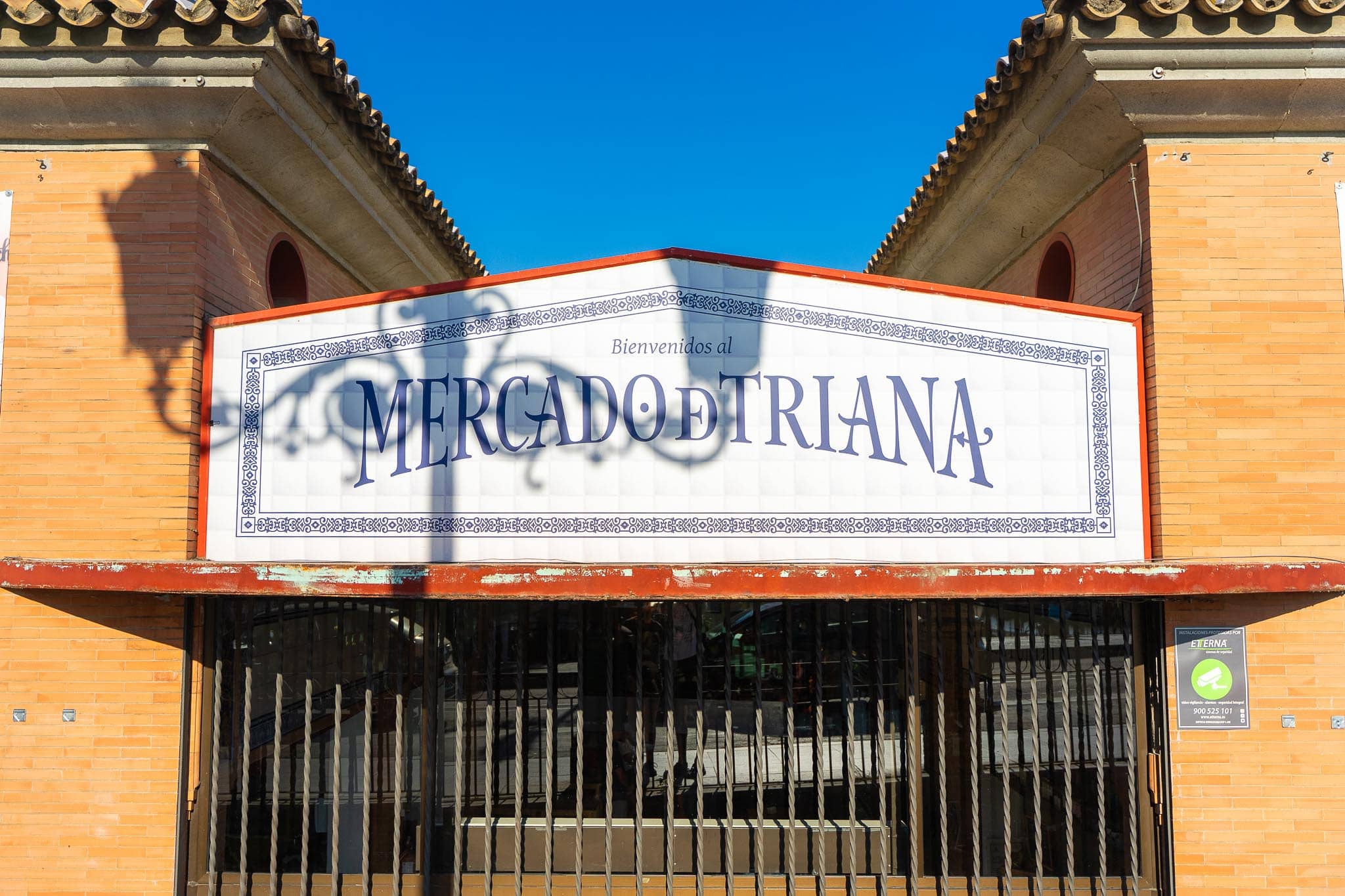 The azulejo sign reading Bienvenidos al Mercado de Triana in blue lettering on white tiles within a decorative tiled border, set into the brick gable above the market's shuttered iron gate, with the shadow of a street lamp thrown across it and a deep blue sky above.