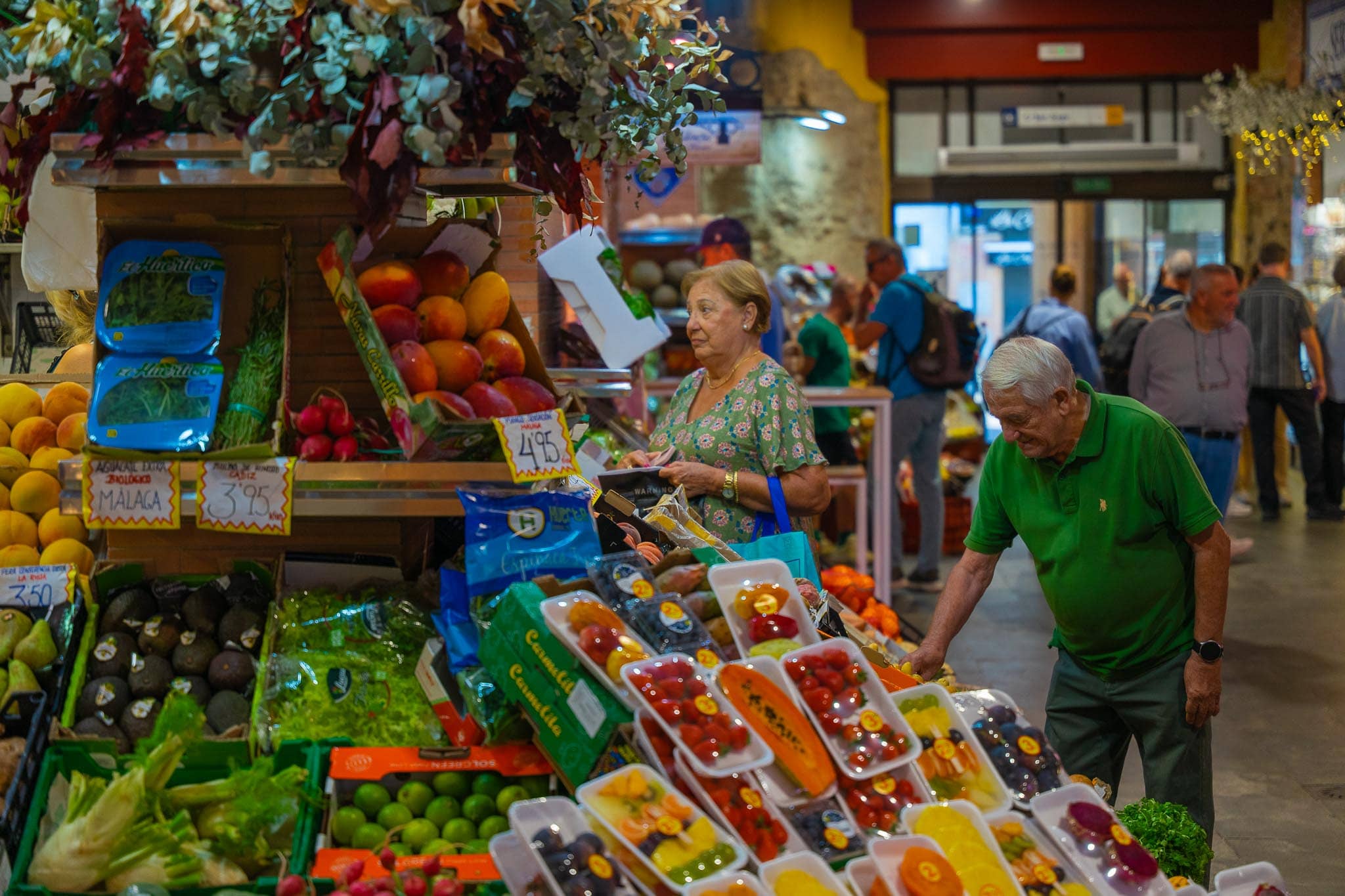 An aisle in the Mercado de Triana mid-morning: a stall of mangos, avocados marked aguacate extra ecológico Málaga 3'95, radishes, fennel and punnets of berries, with two shoppers of retirement age standing at the counter and further stalls and shoppers beyond.
