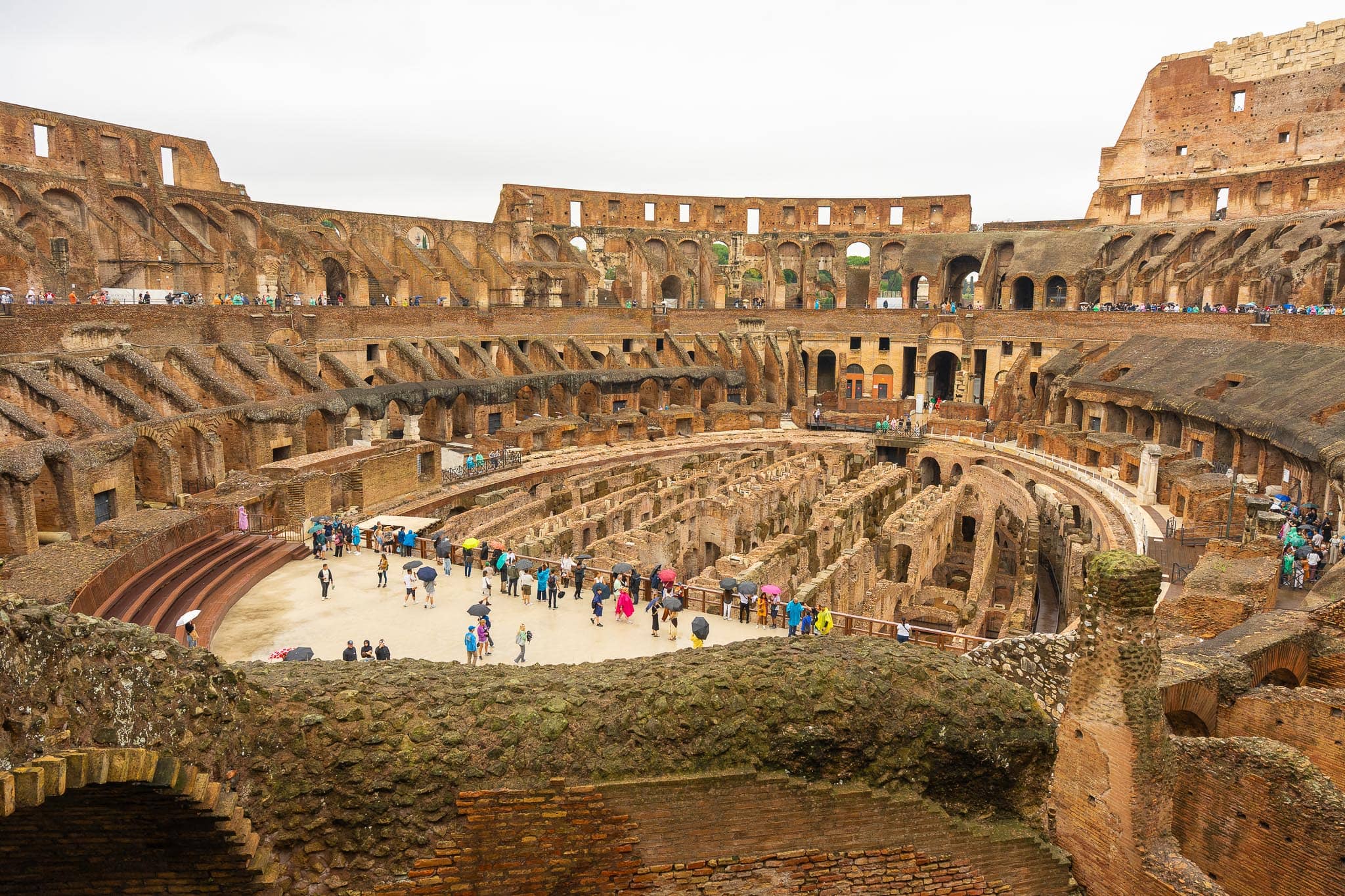 The interior of the Colosseum in Rome: the stripped brick tiers, the exposed corridors of the hypogeum below the arena floor, and visitors on the walkway.