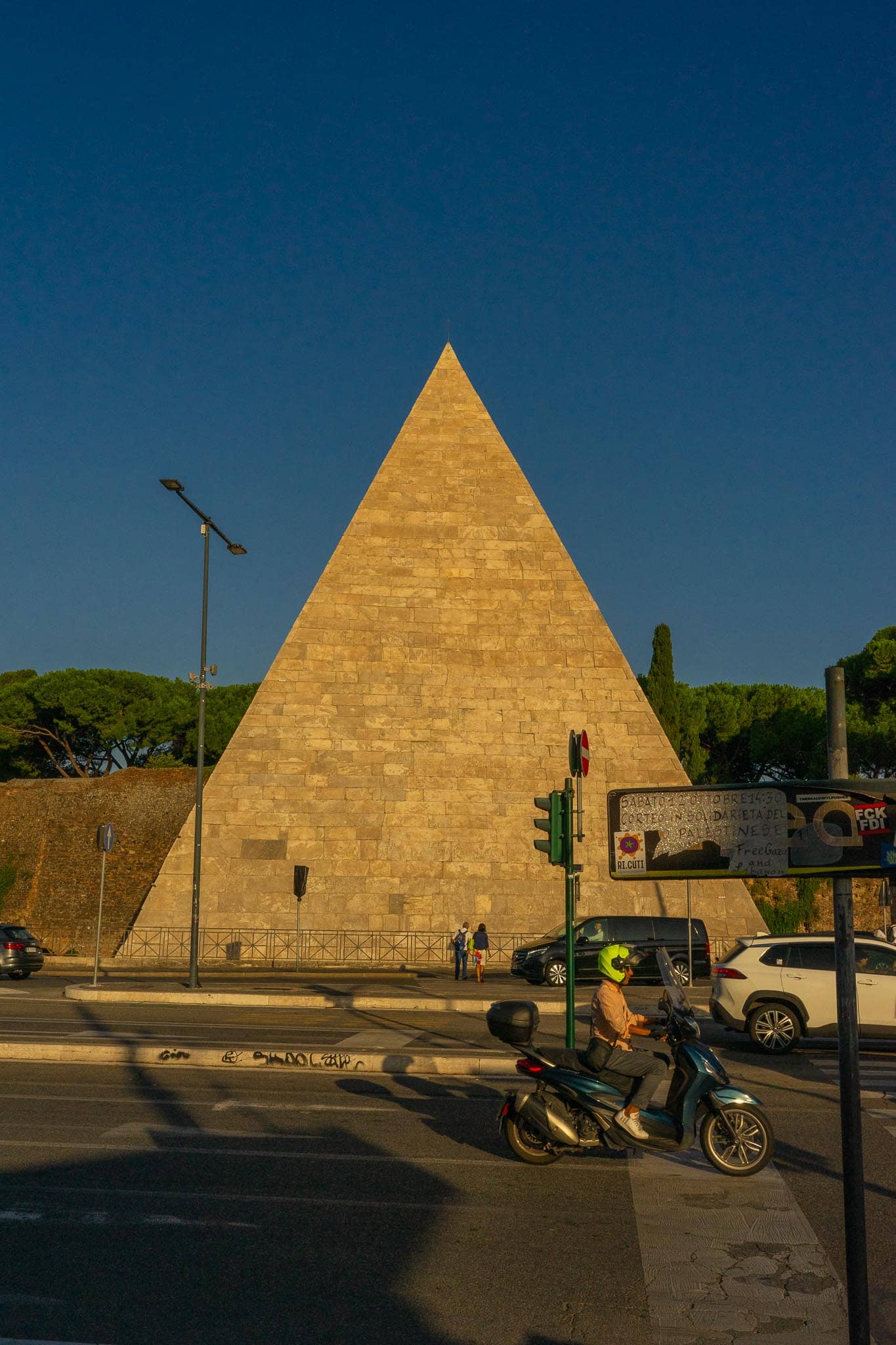 The white marble Pyramid of Cestius in Rome rising behind a road with traffic, a scooter and traffic lights in the foreground under a deep blue sky.