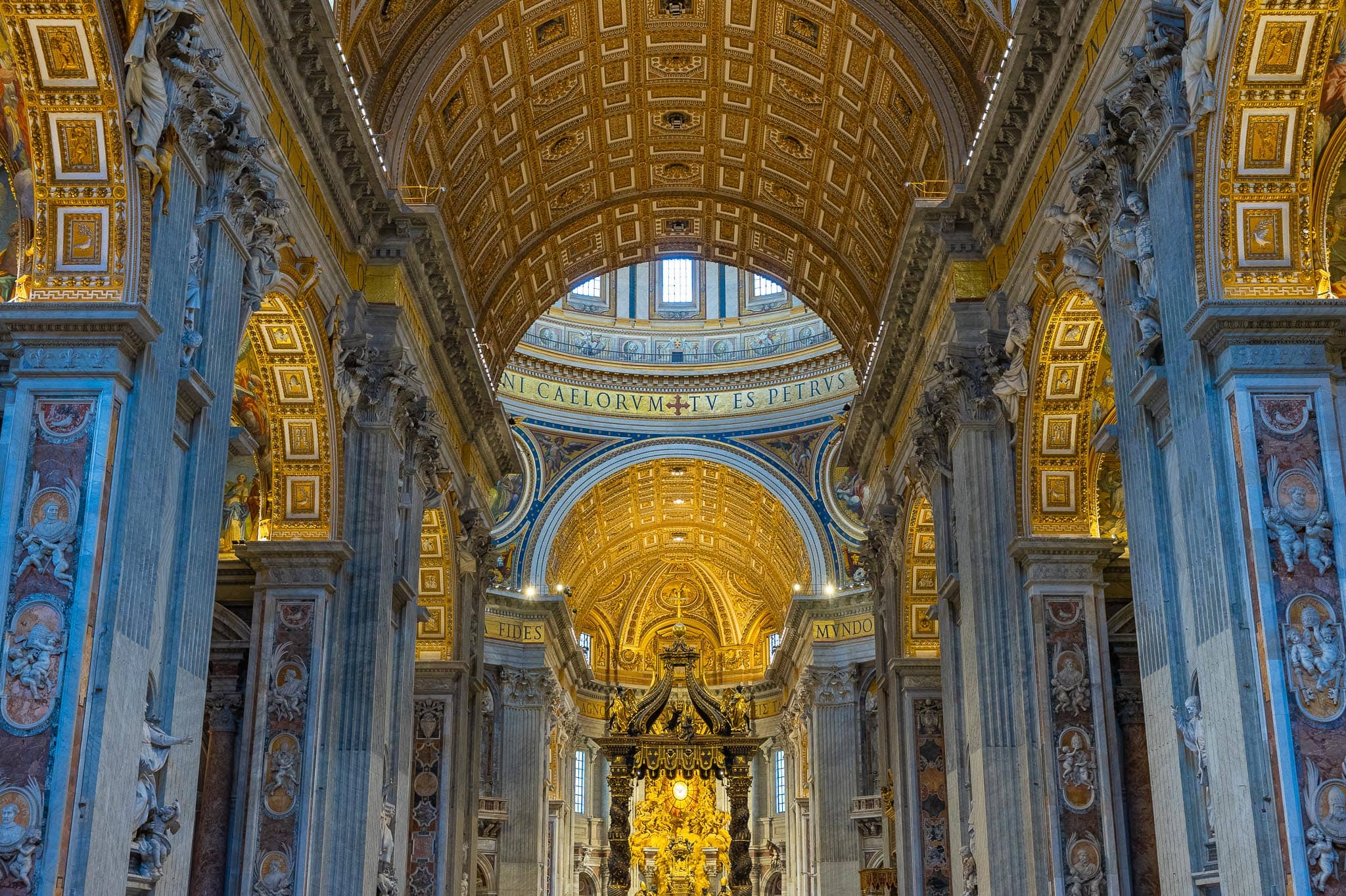 The interior of St Peter's Basilica looking down the nave: a coffered gilt barrel vault, blue-grey marble piers and the bronze baldachin over the high altar.