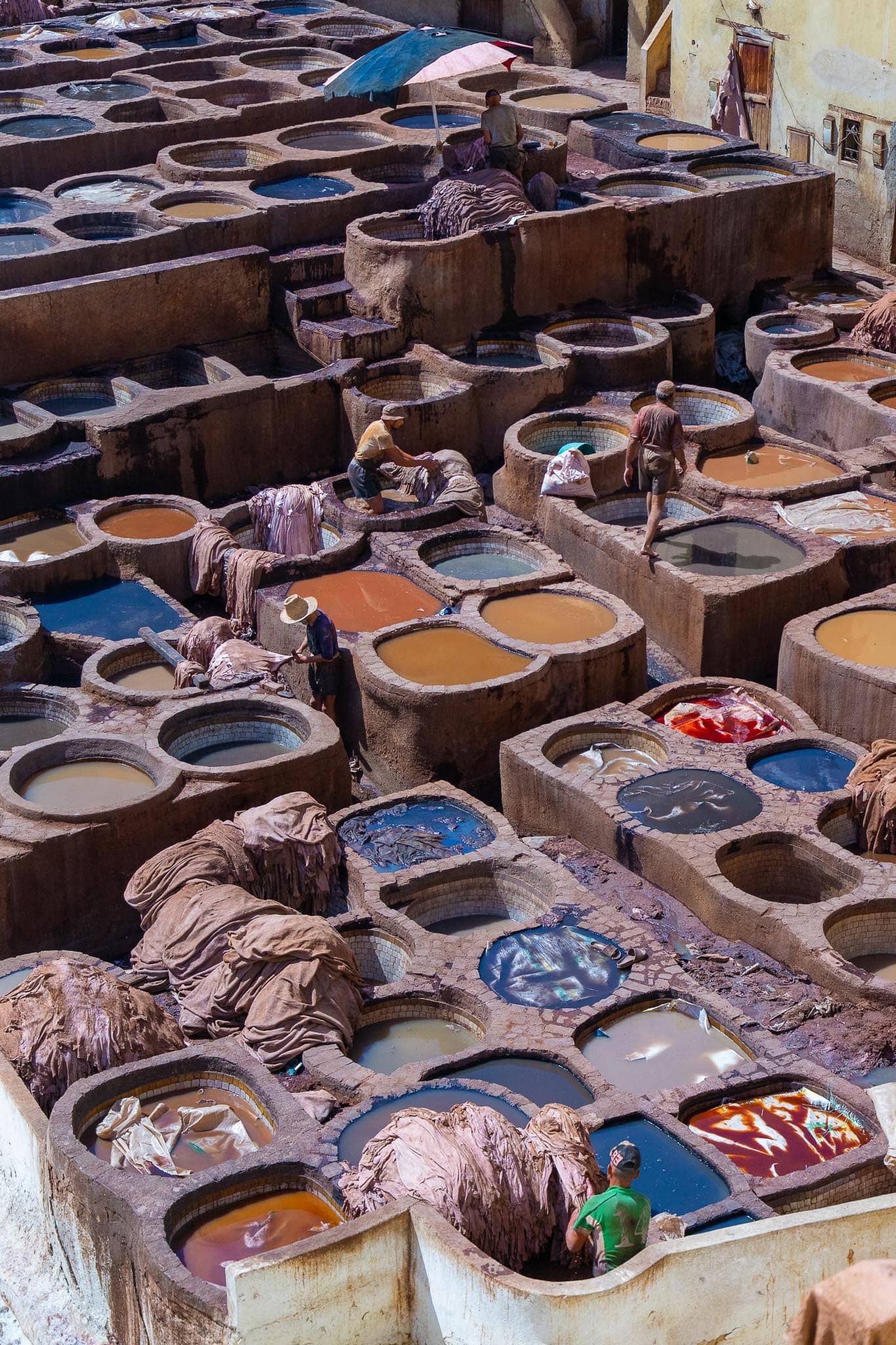 Round stone dye vats at a Fez tannery packed close together, holding red, blue, brown and yellow liquid, with wet hides draped over the rims and a worker among them.