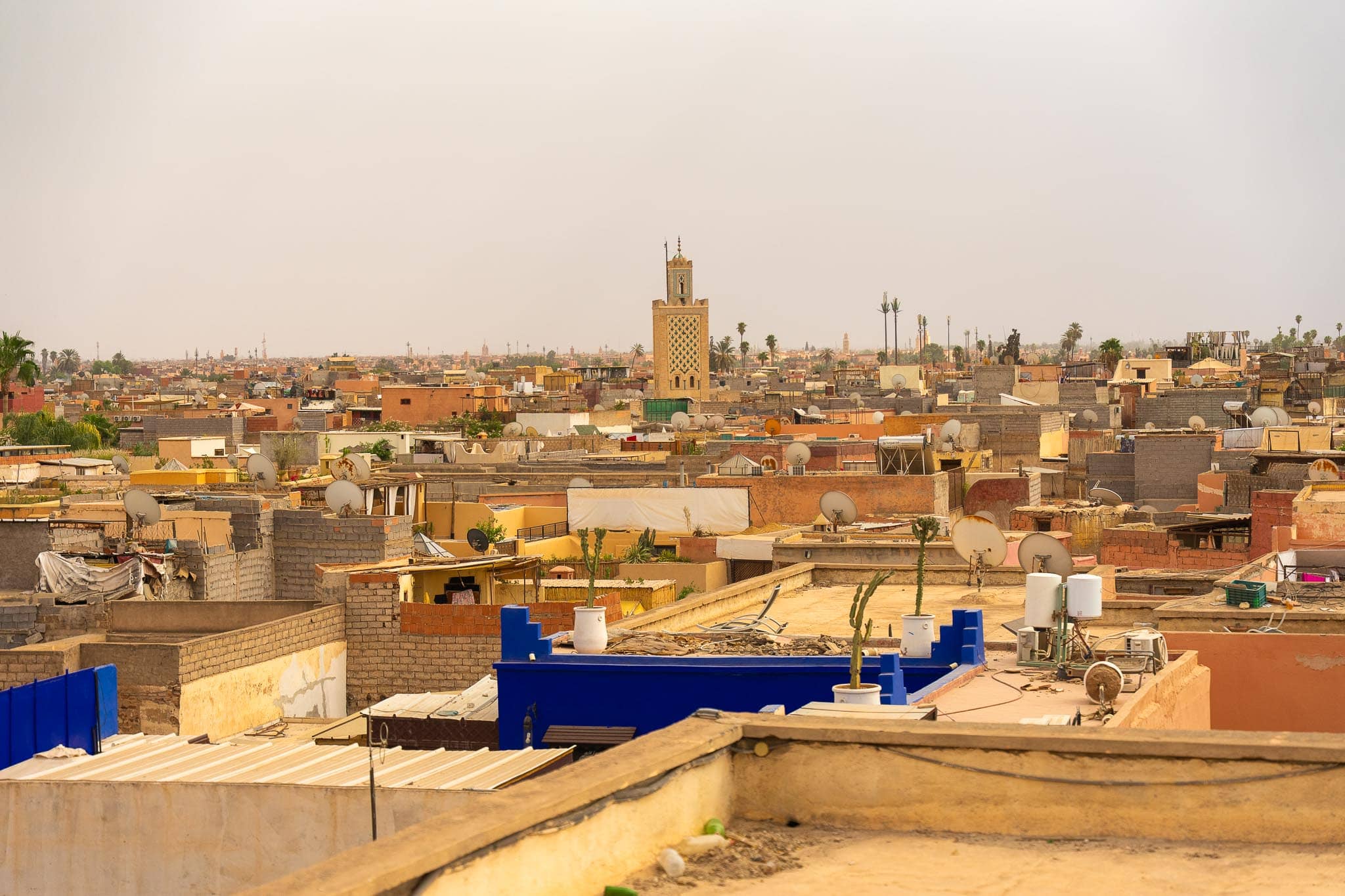 The rooftops of the Marrakech medina in flat afternoon light: pink-brown terraces, satellite dishes and water tanks, with a minaret rising in the middle distance.