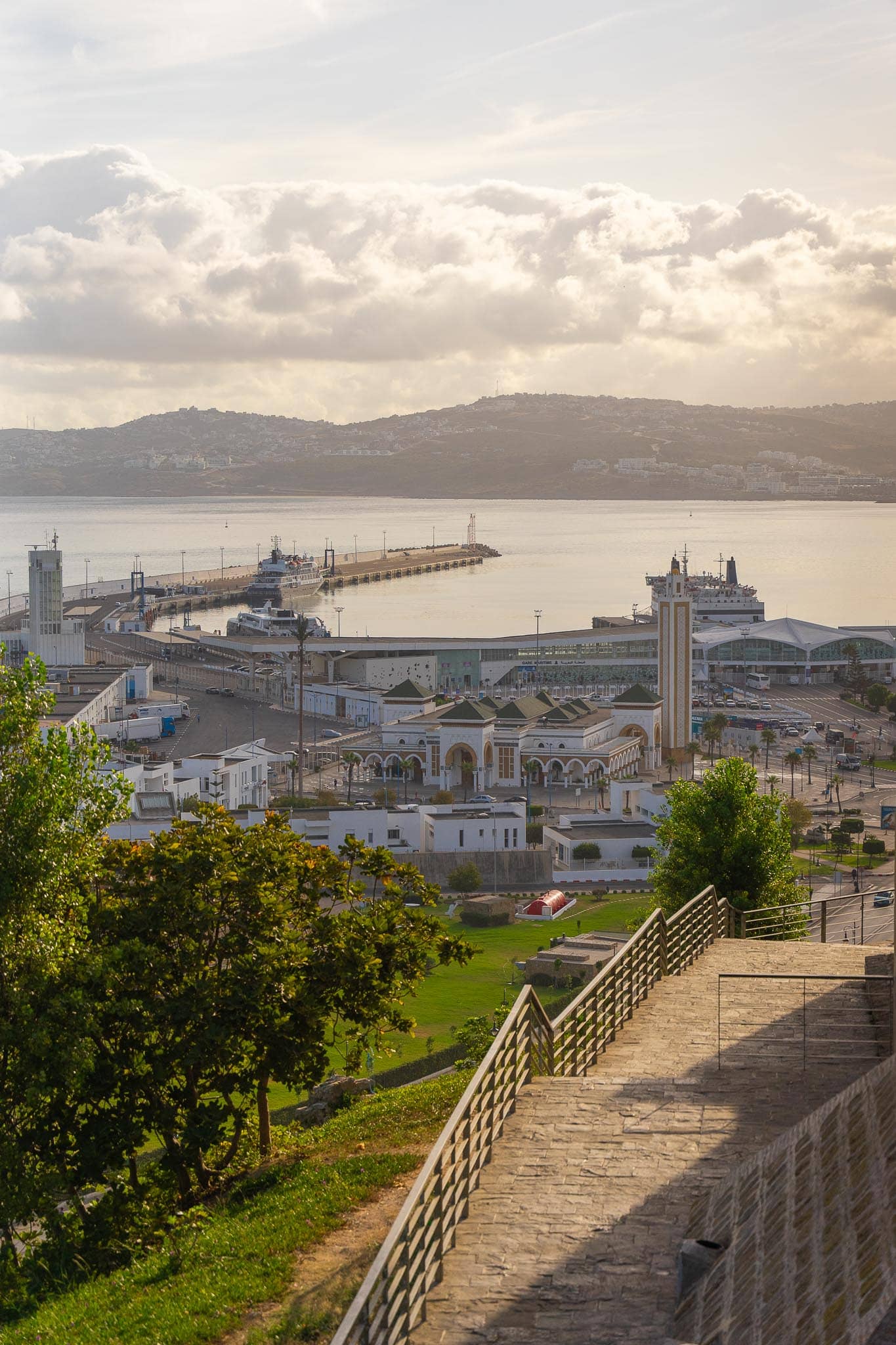 Tangier from a wooded hillside above: white apartment blocks, the port with its jetties and moored ships, and the hazy coastline curving away across the bay.